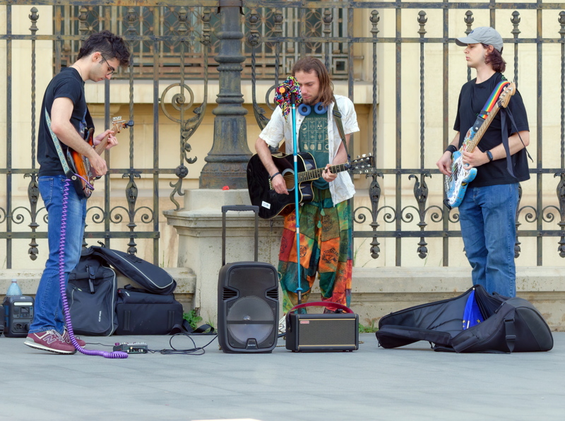 A trio of street musicians playing acoustic and electric guitars in front of a decorative iron fence